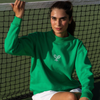 Woman in a Kelly Green CourtLife Tennis Mock Neck sweatshirt with white crossed racquet embroidery sitting against a tennis net