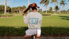Back of a woman on a golf course in an Ivory CourtLife Happiest on the Course golf sweatshirt with blue lettering