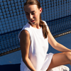 Woman in an Ivory CourtLife Pique Tank and white skort sitting against the net on a blue tennis court